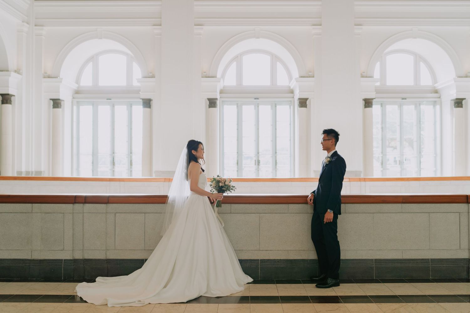 Bride and groom standing apart facing each other before arched colonial building windows.