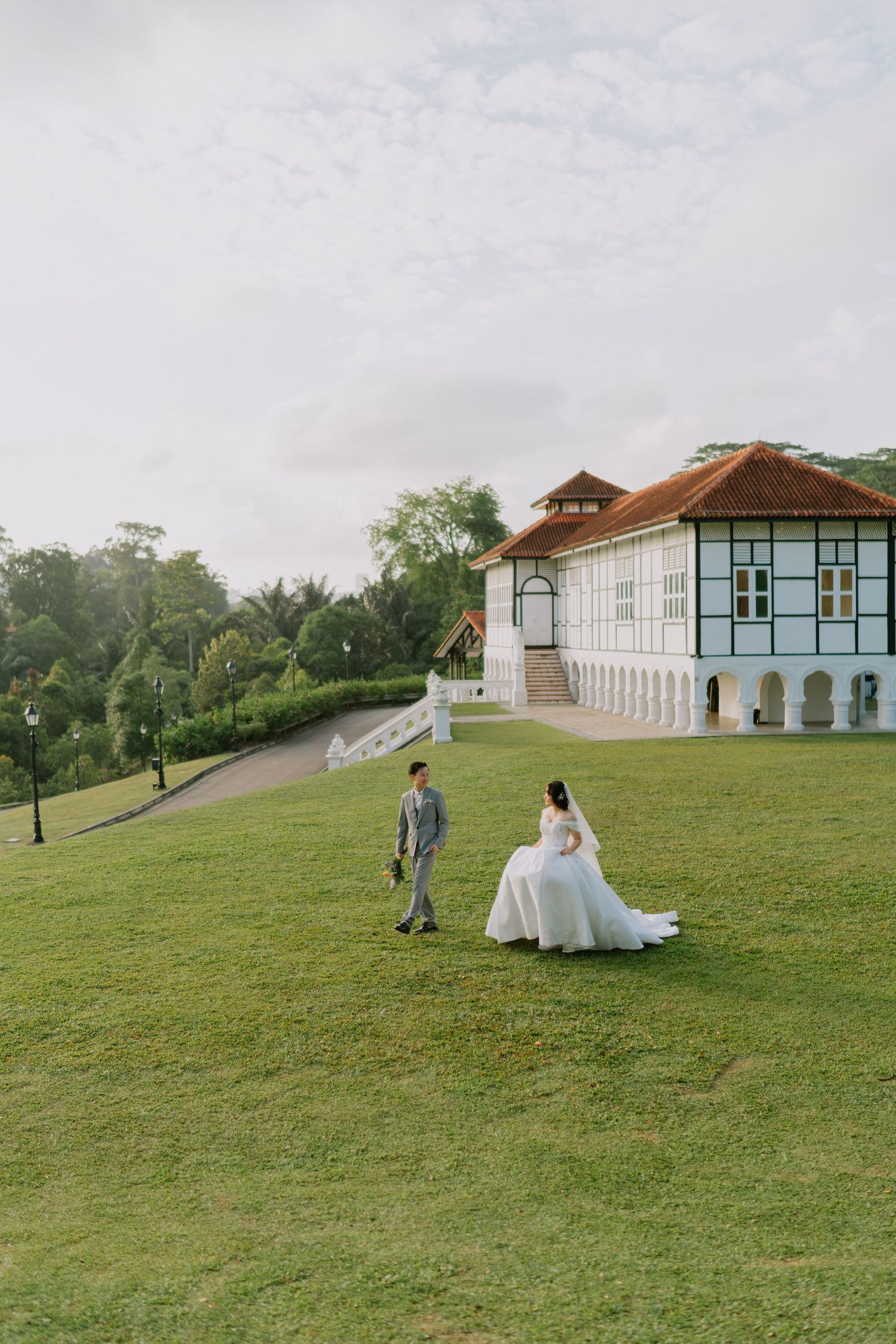 Bride and groom walk on green lawn before a colonial blue and white building on their wedding day.