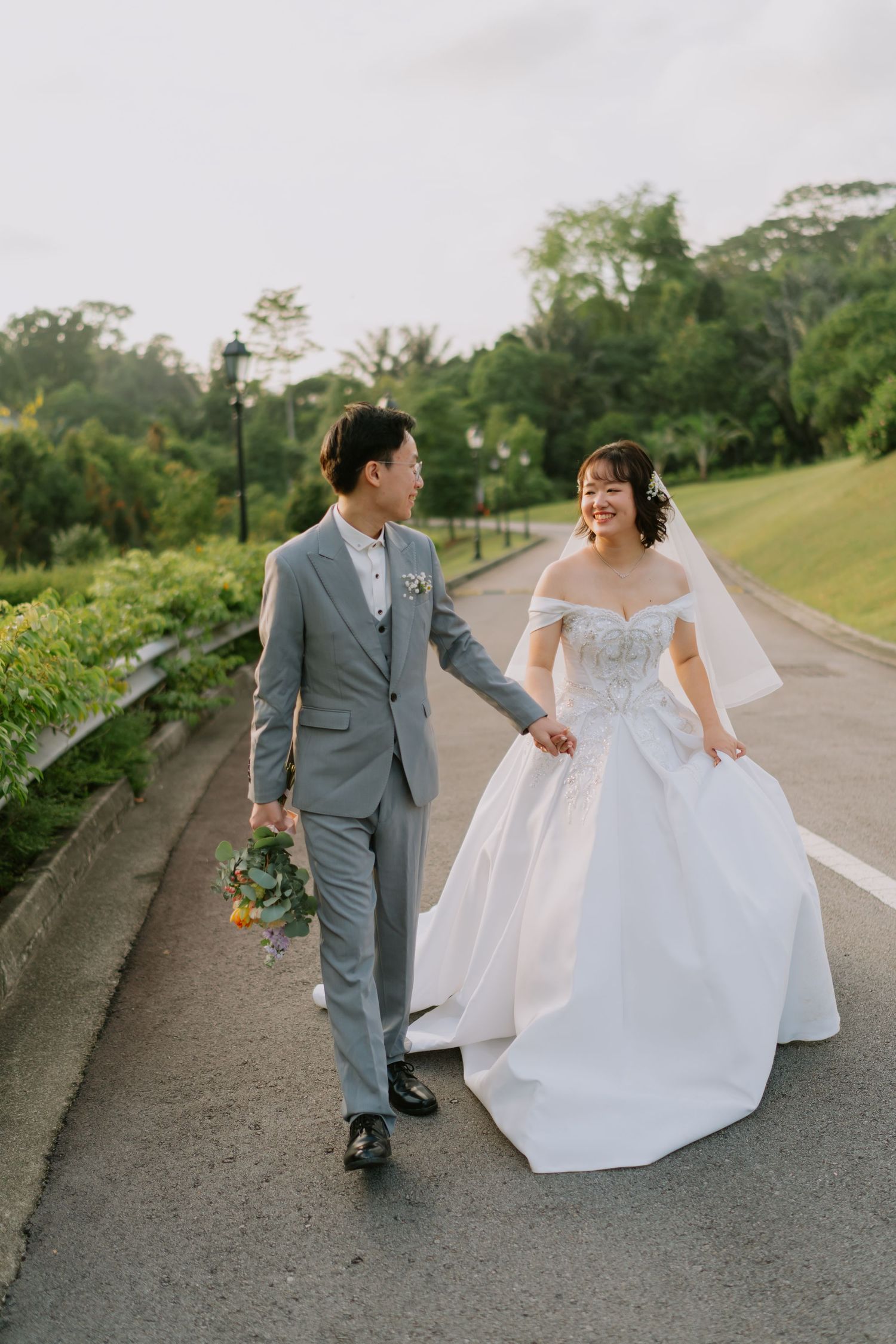 Smiling Asian bride and groom walk hand-in-hand on road, she in white ballgown, he in gray suit.