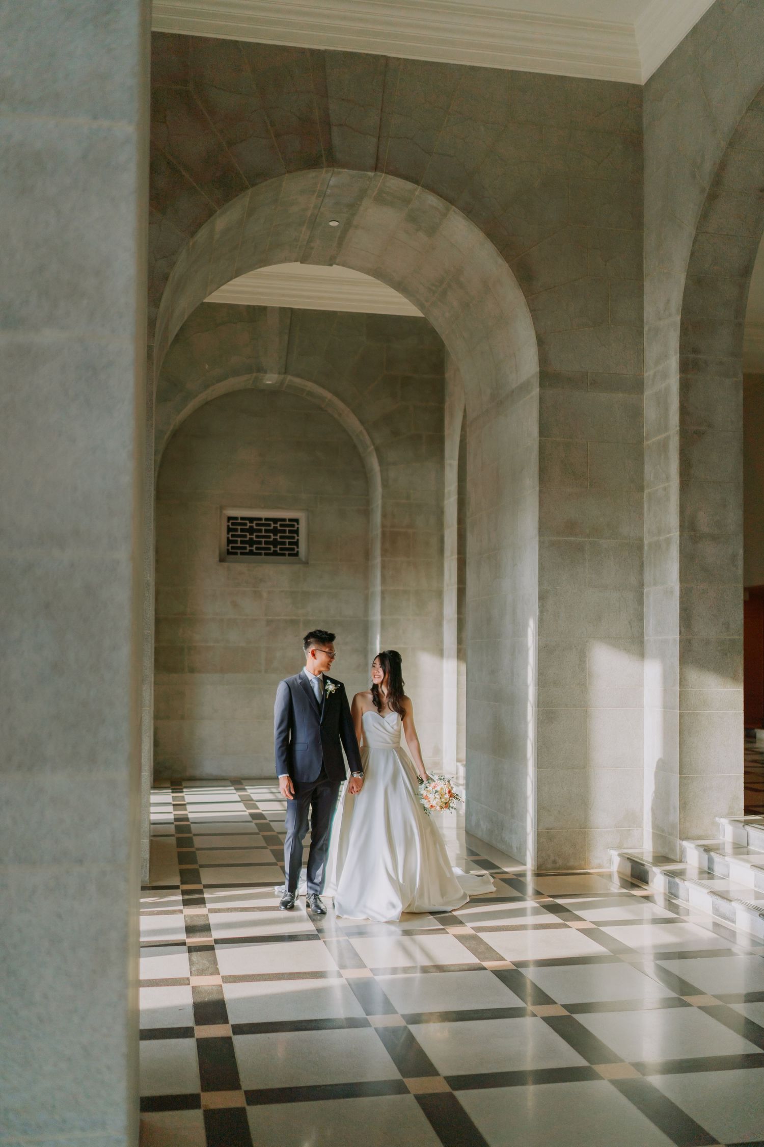 Bride and groom walk hand in hand through elegant stone arched corridor with checkered marble floor.