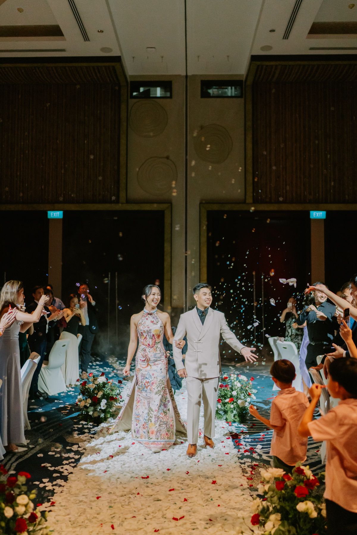 Bride and groom walk down petal-lined aisle as guests toss confetti at elegant indoor wedding reception.