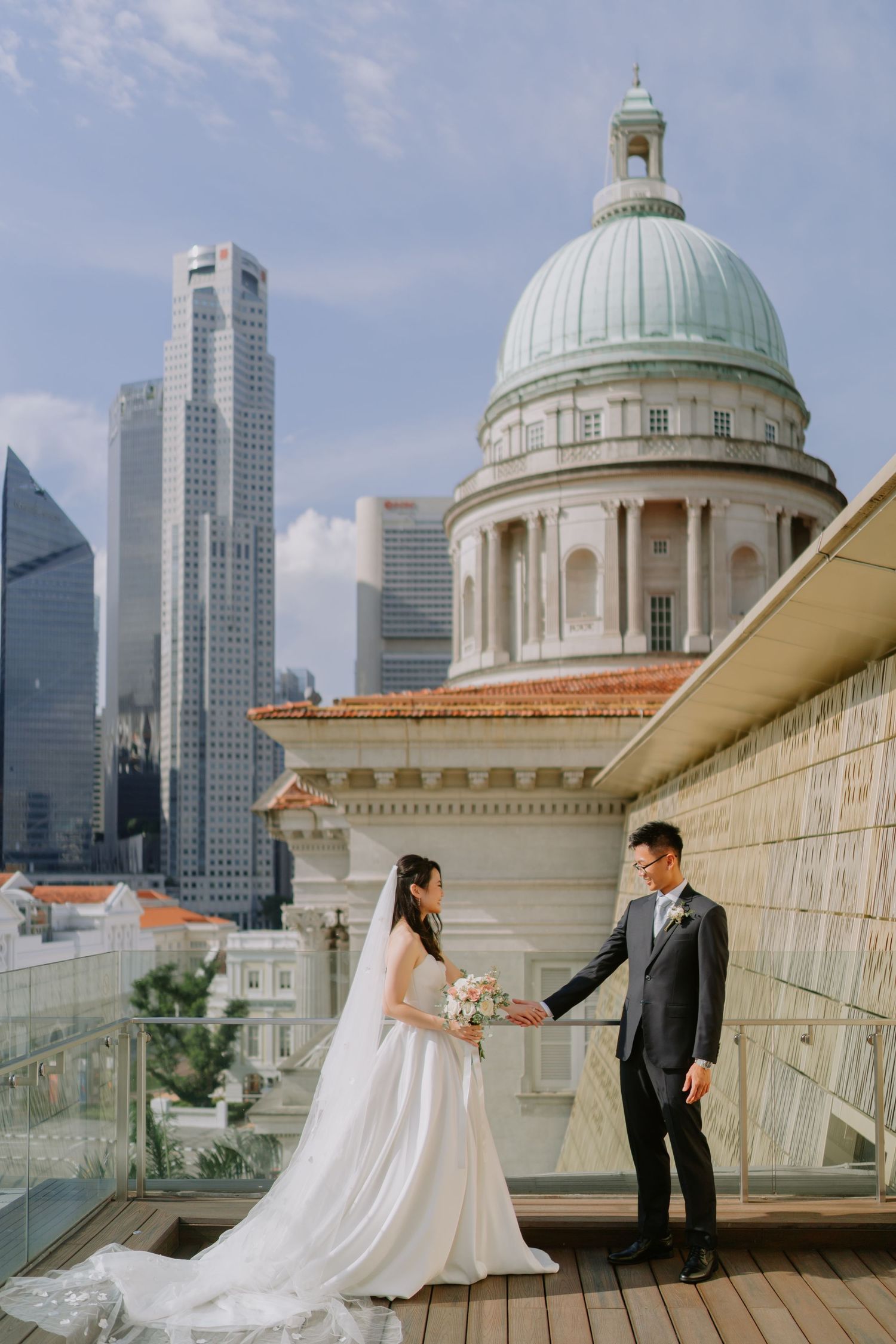 Bride and groom hold hands on rooftop terrace with Singapore skyline and historic domed building backdrop.