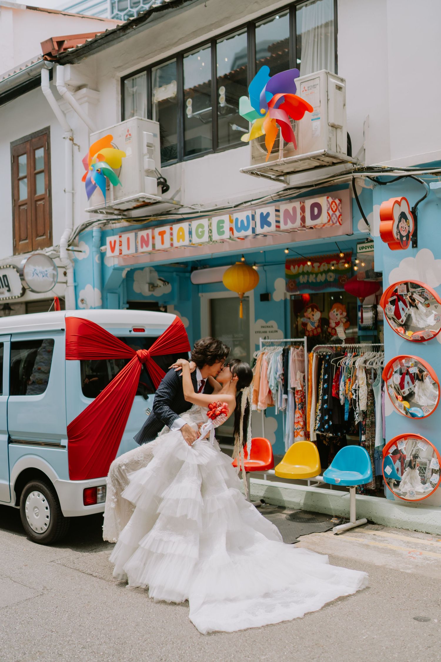 Groom dipping bride in wedding gown outside vibrant Vintage Weekend shophouse with blue van.