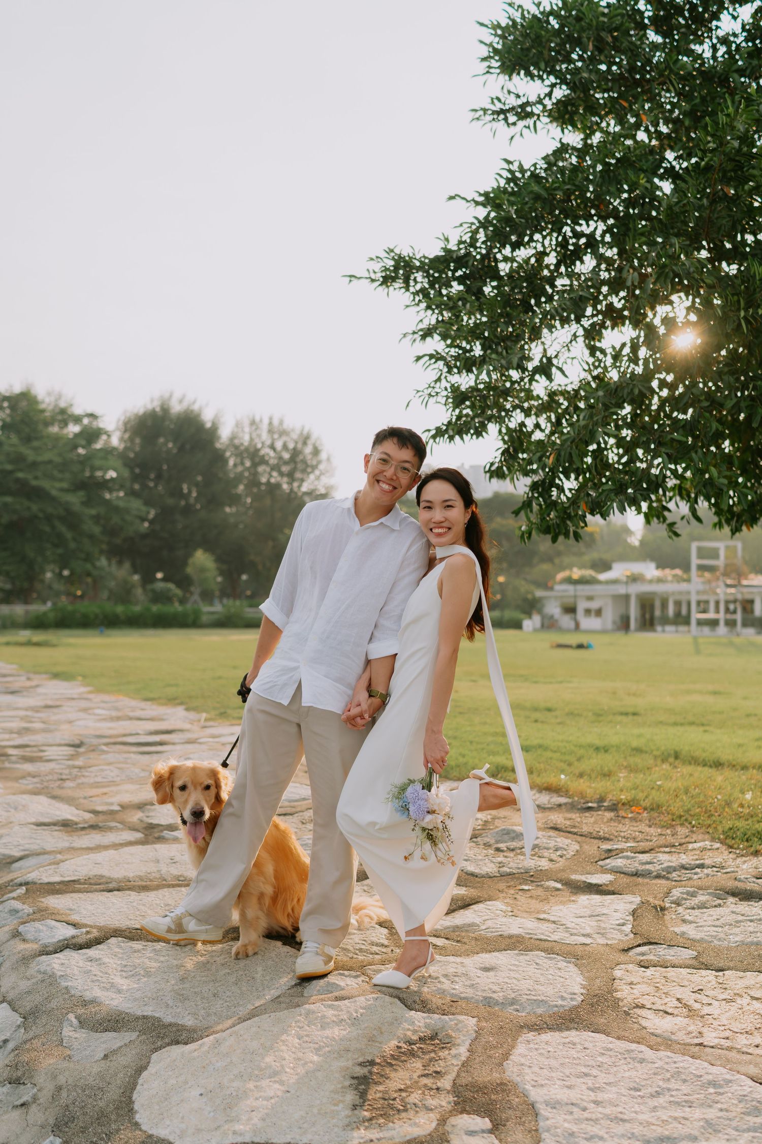 Smiling couple in white outfits walking golden retriever on stone path under tree.