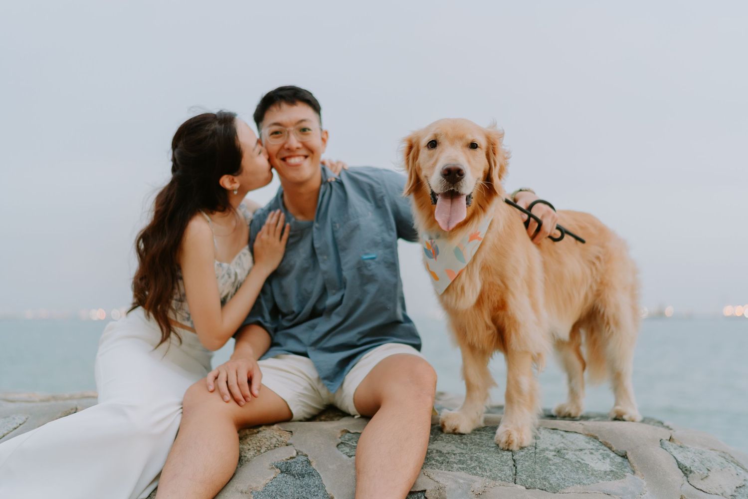 Smiling couple sitting on rocks by the sea with golden retriever wearing a bandana.