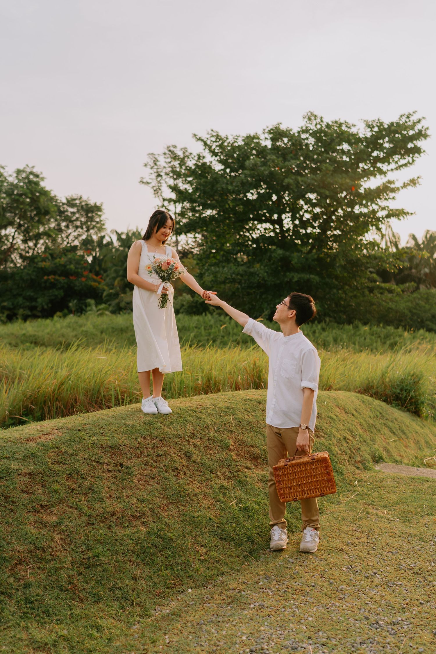 Man reaching up to hold woman's hand on grassy hill at sunset, both carrying picnic items.