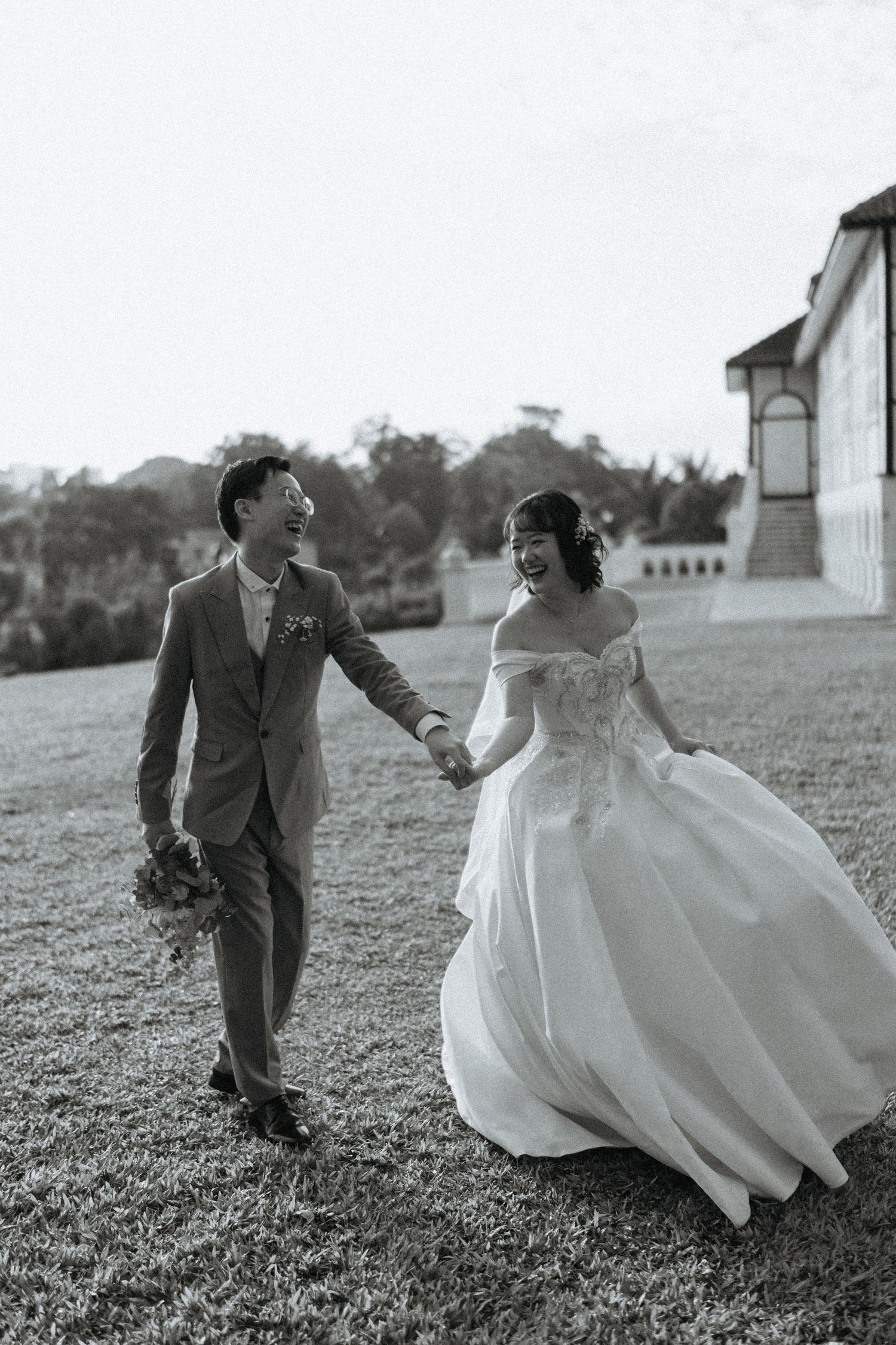 Black and white photo of laughing Asian bride and groom holding hands while walking on grass.