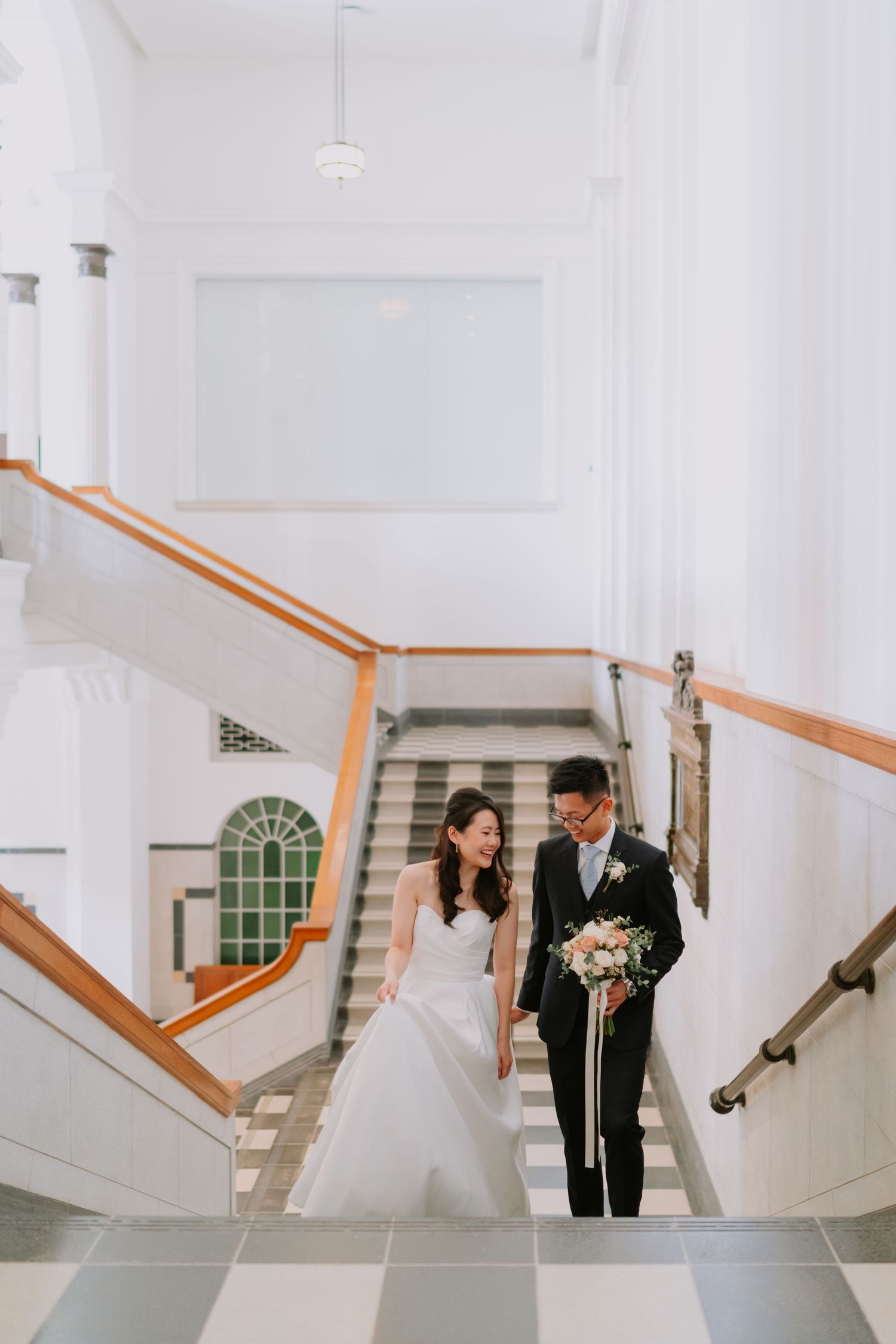 Smiling bride and groom walking hand in hand down bright white colonial building staircase.