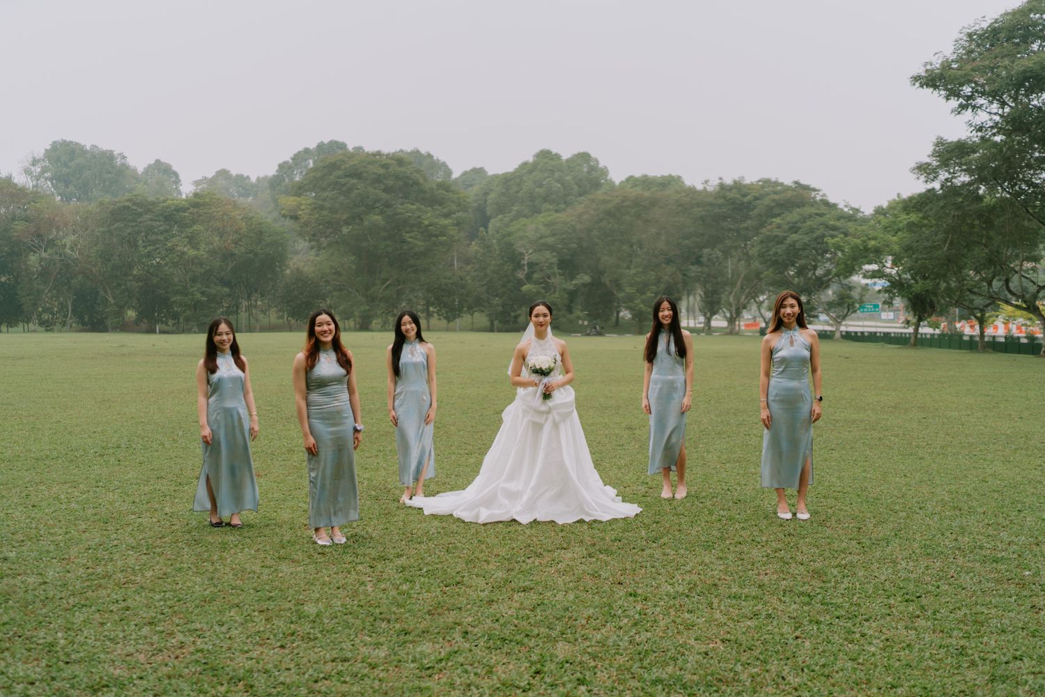 Bride in white gown poses with six bridesmaids in sage green dresses on lush green lawn surrounded by trees.
