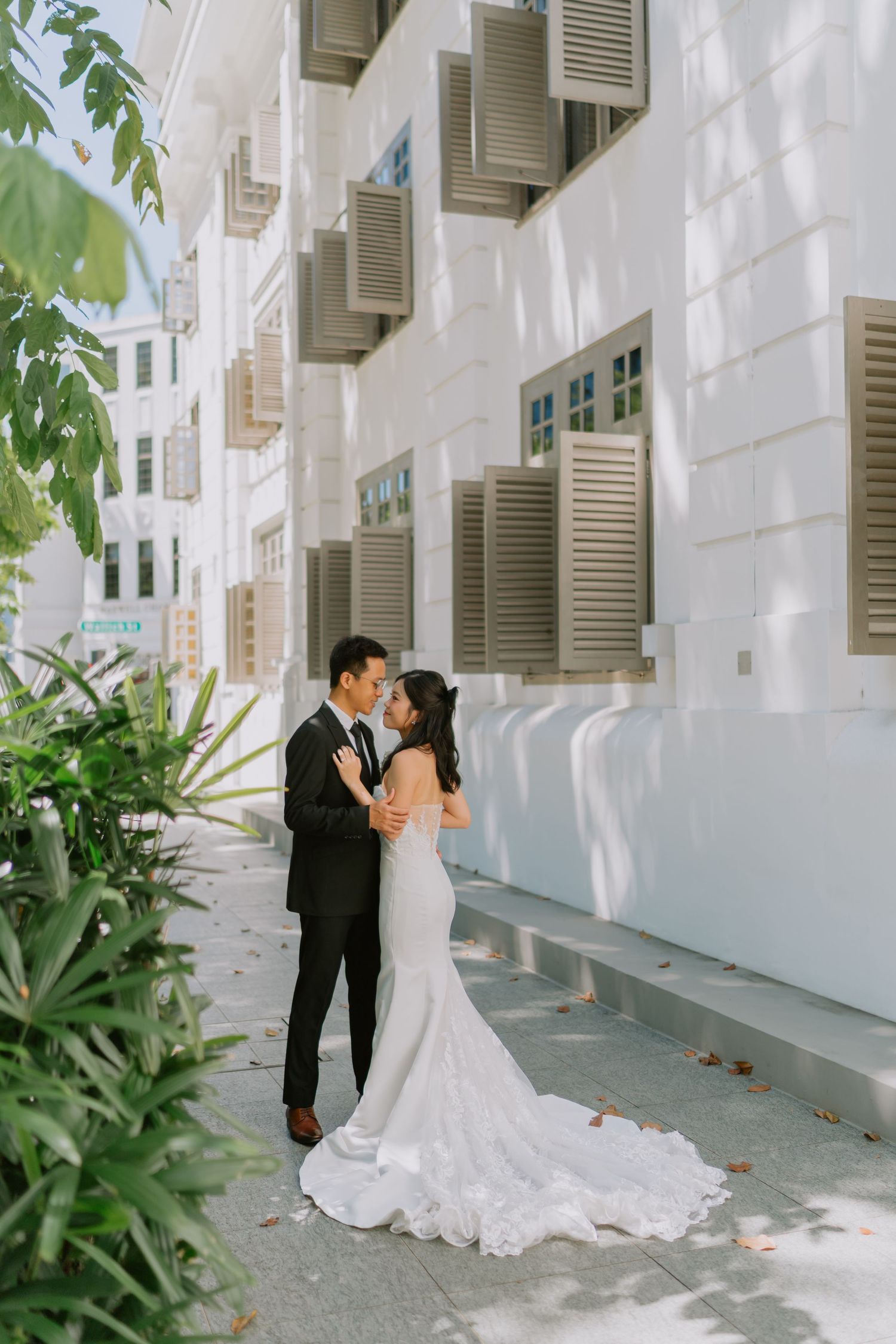 Asian bride and groom sharing romantic moment beside white colonial building with shuttered windows.