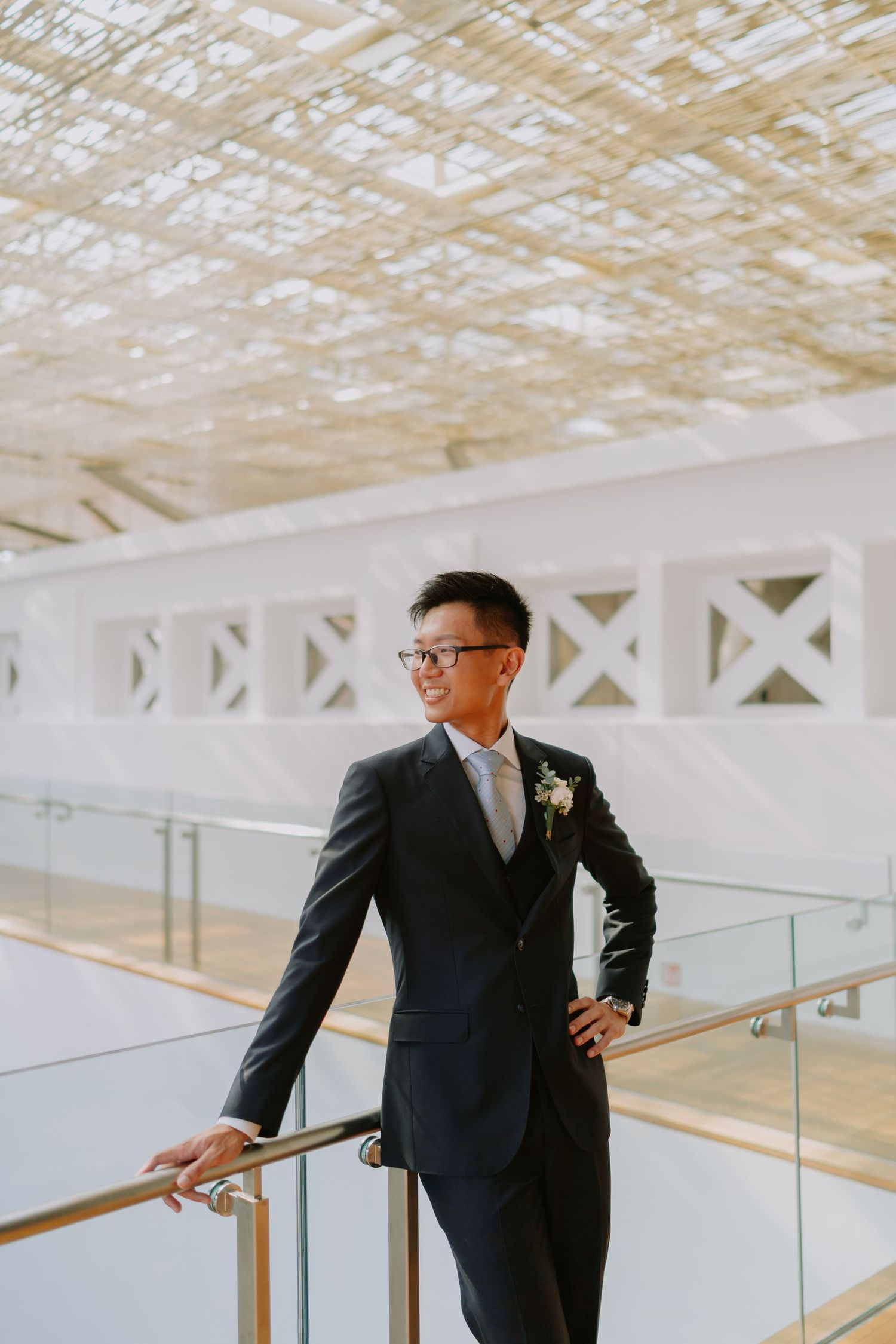 Dapper Asian groom in dark suit with boutonniere leans on glass railing in modern venue.