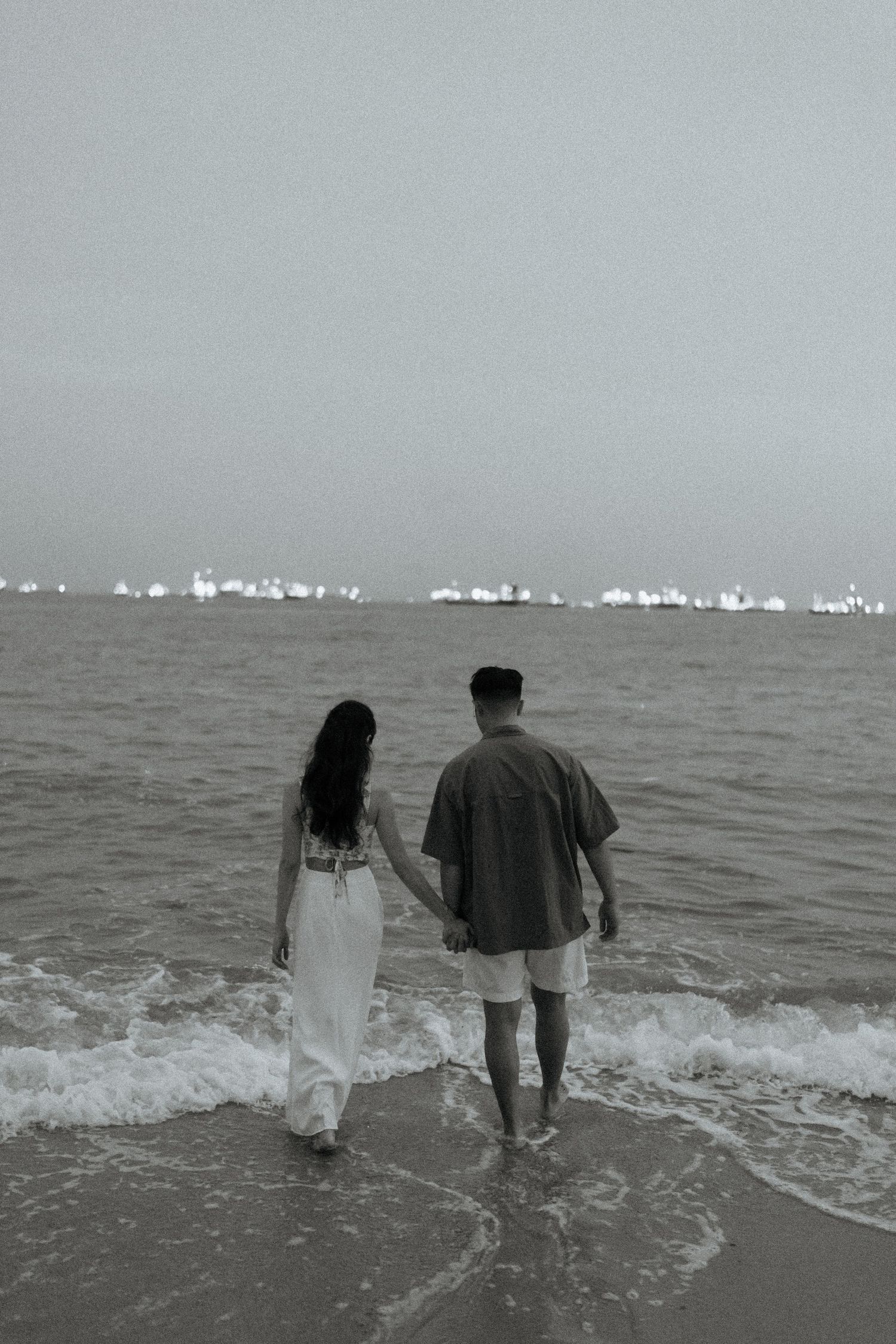 Black and white photo of a couple holding hands while walking along the ocean shoreline.