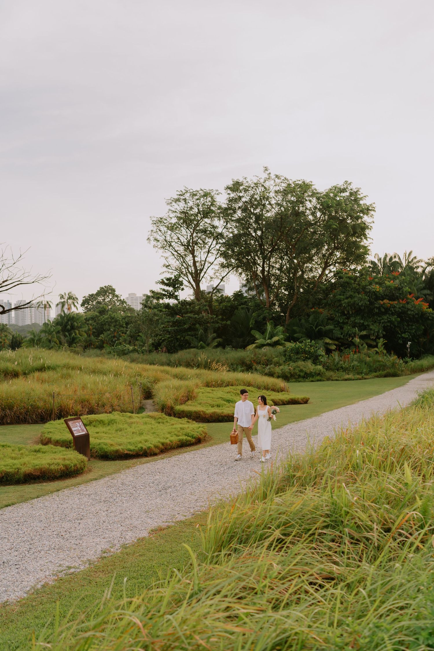 Couple walking hand-in-hand along a scenic gravel path through lush green gardens at dusk.