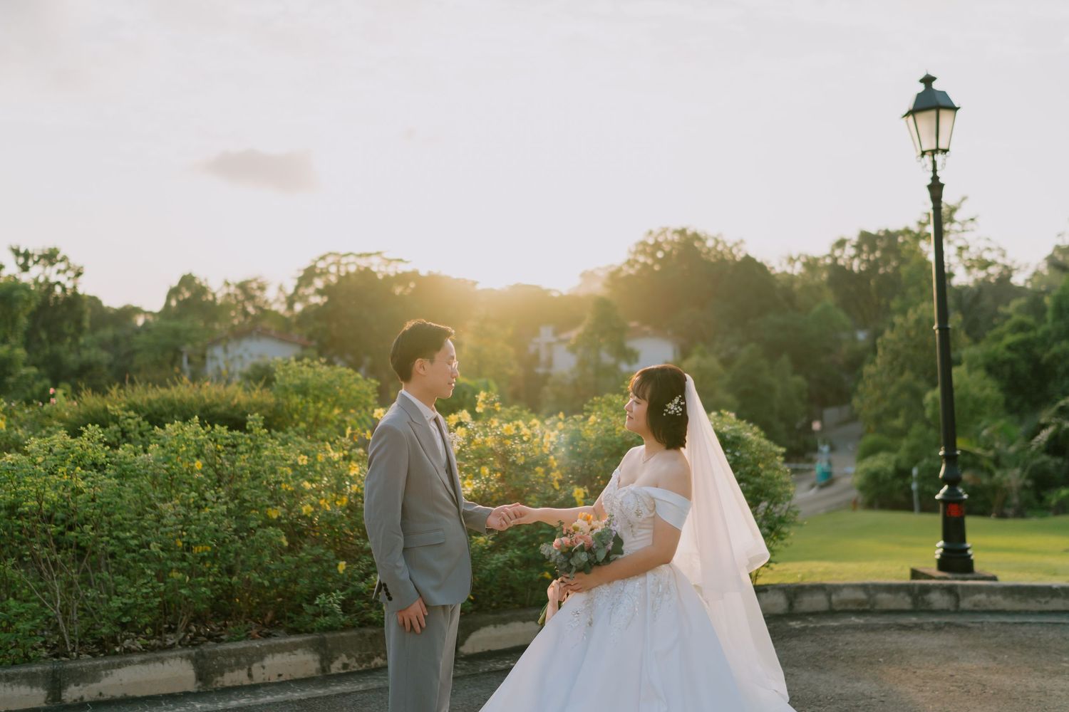 Newlywed couple holding hands at golden hour sunset in a scenic garden with lamp post nearby.