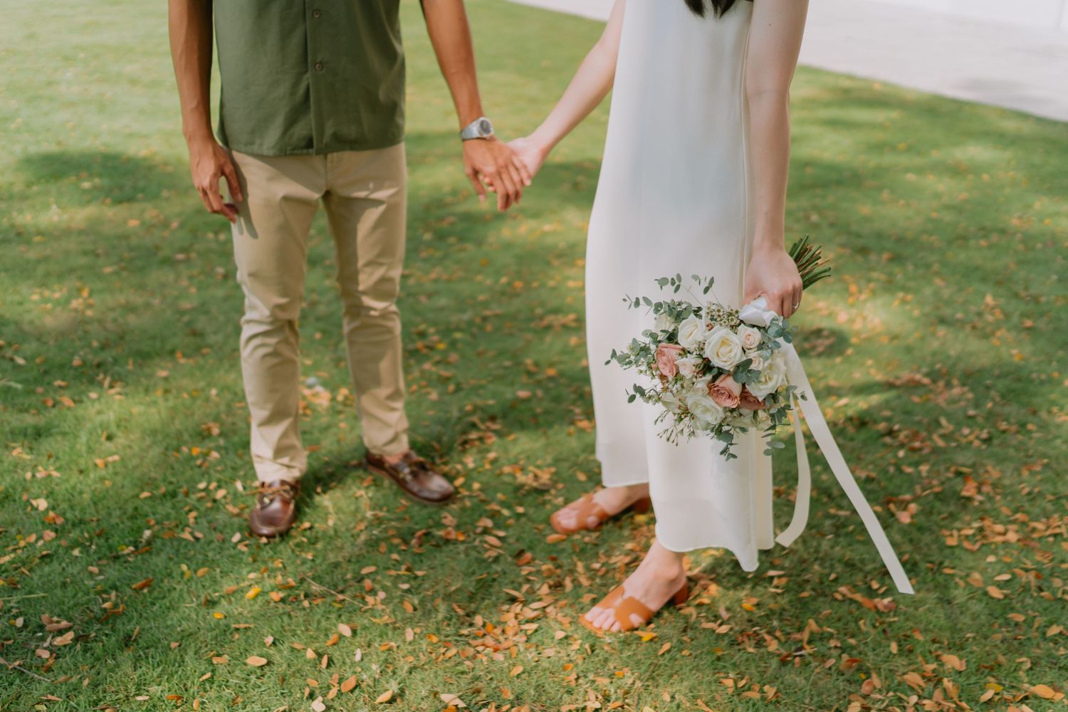 Close-up of couple holding hands on grass, bride carrying soft pink and white bouquet.