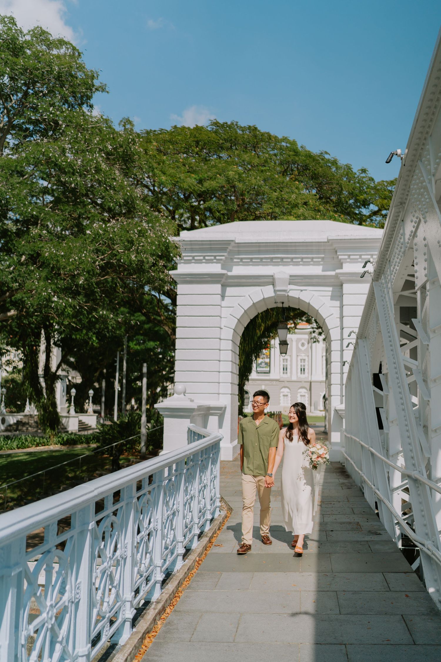 Couple holding hands walking on a white ornate bridge under blue sky.