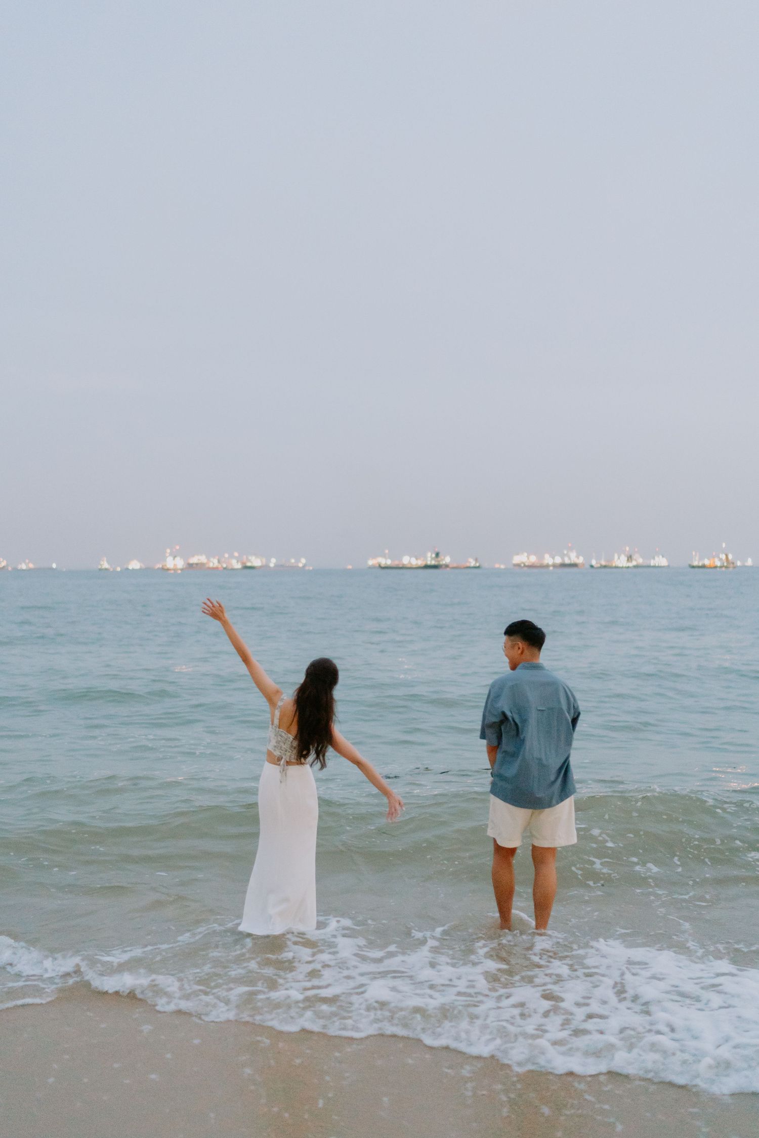 Couple standing in ocean waves at the beach, woman raising her arm joyfully toward the sea.