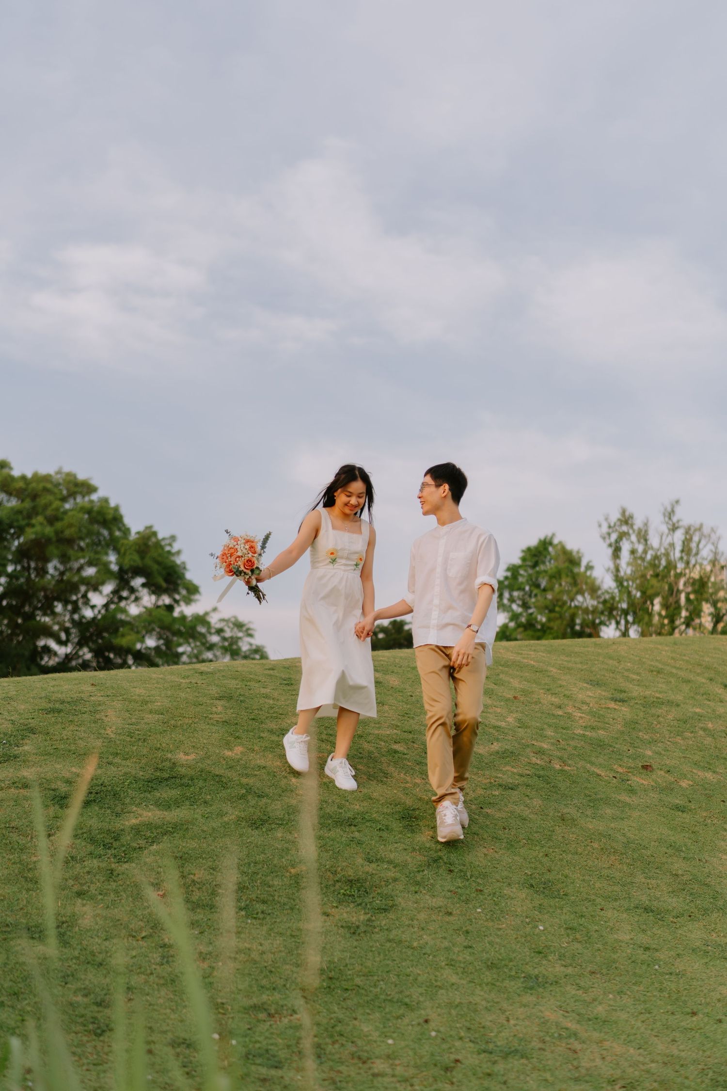 A joyful couple runs hand in hand on a grassy hill, the woman in a white dress holding a bouquet.