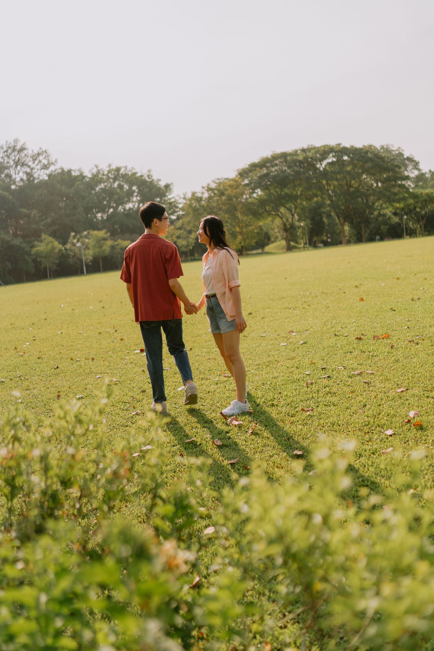 A couple walks hand in hand across a sunlit green field surrounded by trees.