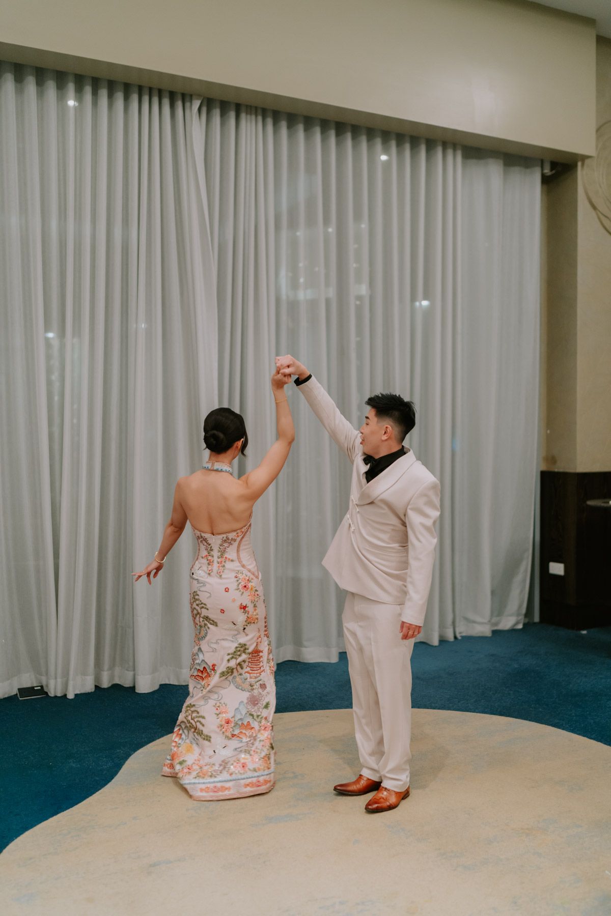 Couple sharing first dance in elegant ballroom, bride in floral gown and groom in white suit holding hands raised.