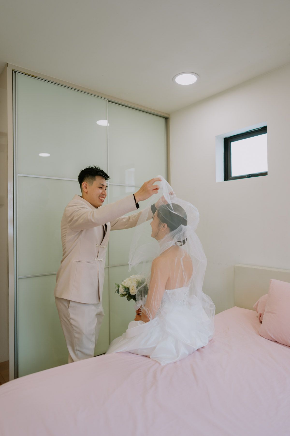 Groom in beige suit placing veil on bride in white wedding dress seated on pink bed in bright modern bedroom.