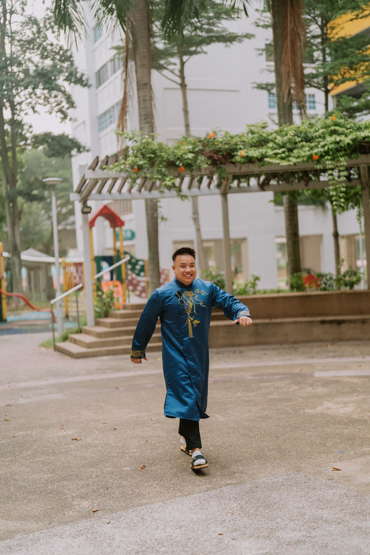 Young man in traditional blue Vietnamese ao dai walks confidently outdoors near a playground and palm trees.