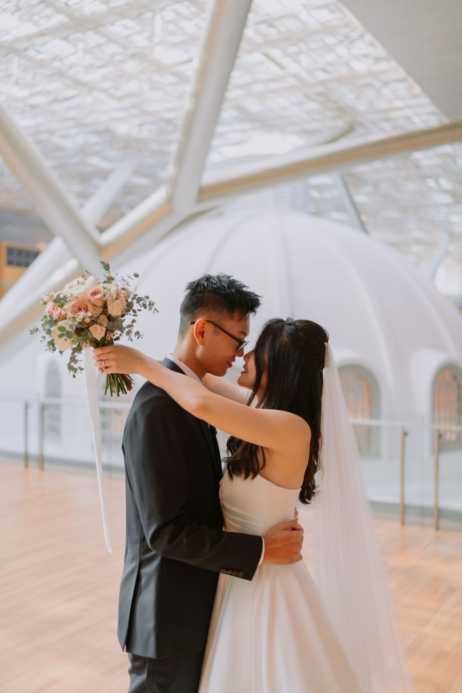 Asian bride and groom sharing intimate nose-to-nose moment, bride holding pink bouquet in modern white venue.