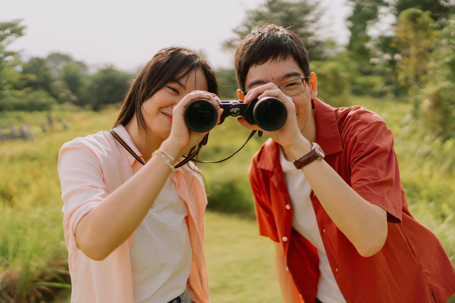 A smiling couple looks through binoculars together outdoors in a green landscape.