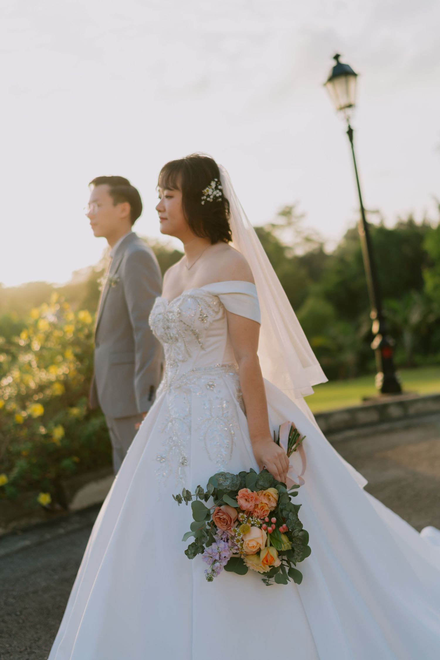 Bride in off-shoulder white gown holds colorful bouquet as groom in gray suit stands behind at golden hour.