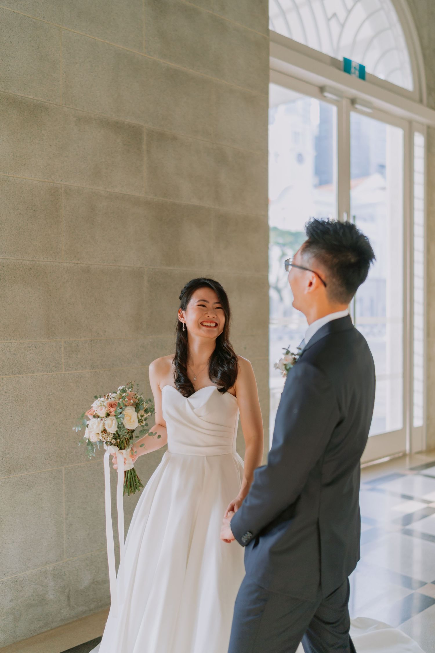 Smiling bride in white gown holds bouquet while facing groom in dark suit by large window.