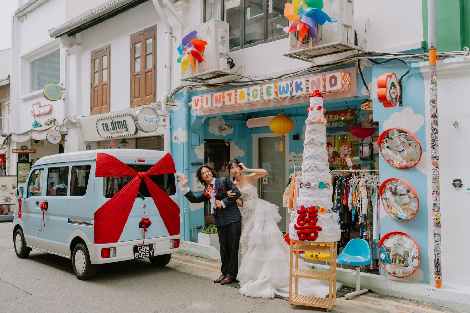 Wedding couple posing beside light blue van with red bow outside colorful Vintage Weekend store.
