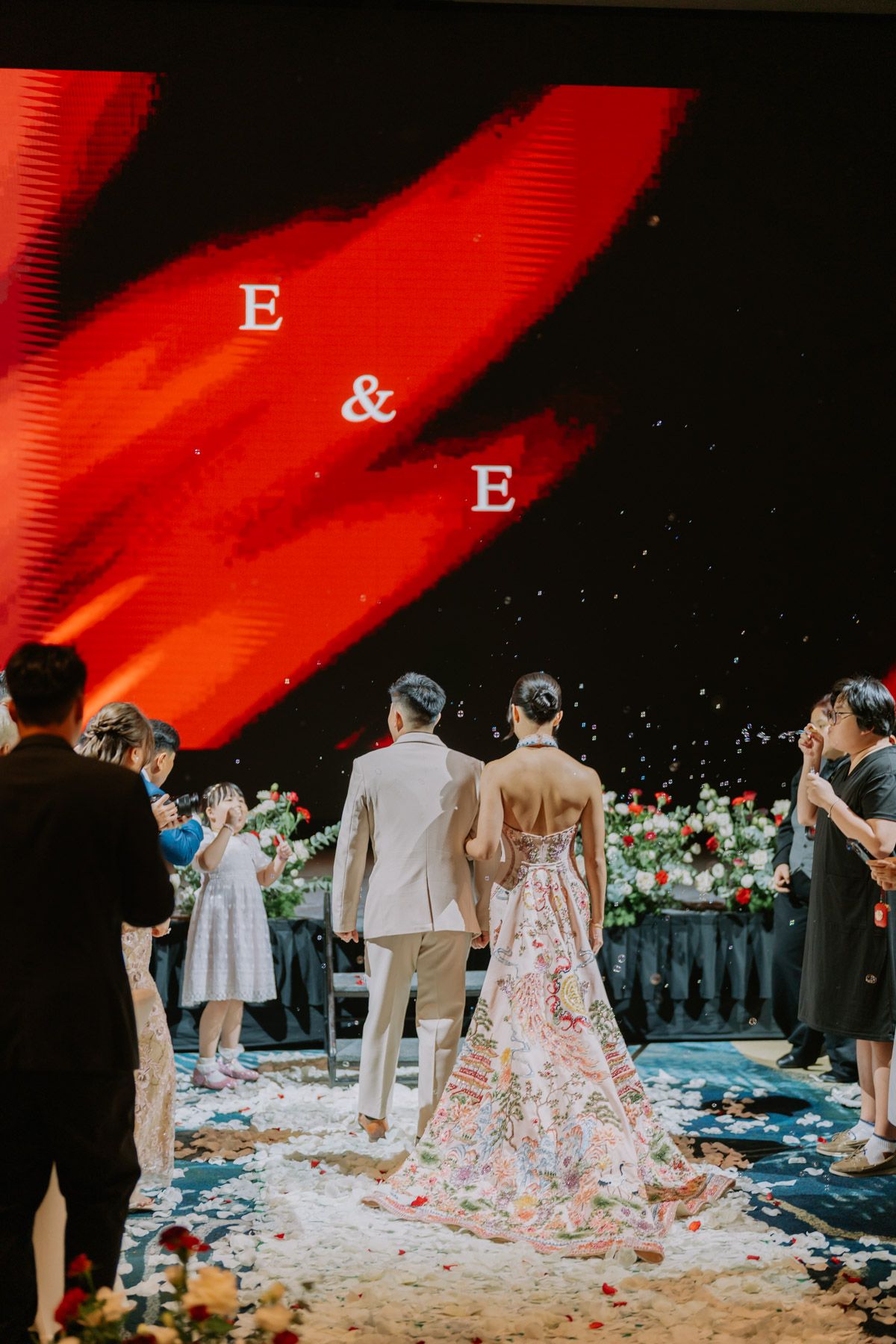 Couple walks down wedding aisle in floral gown and suit, guests toss petals, red E&E monogram display backdrop.