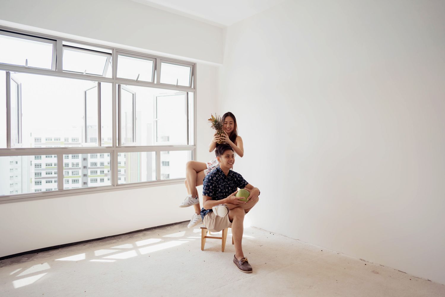 Couple poses playfully in empty white room with large windows and city view.