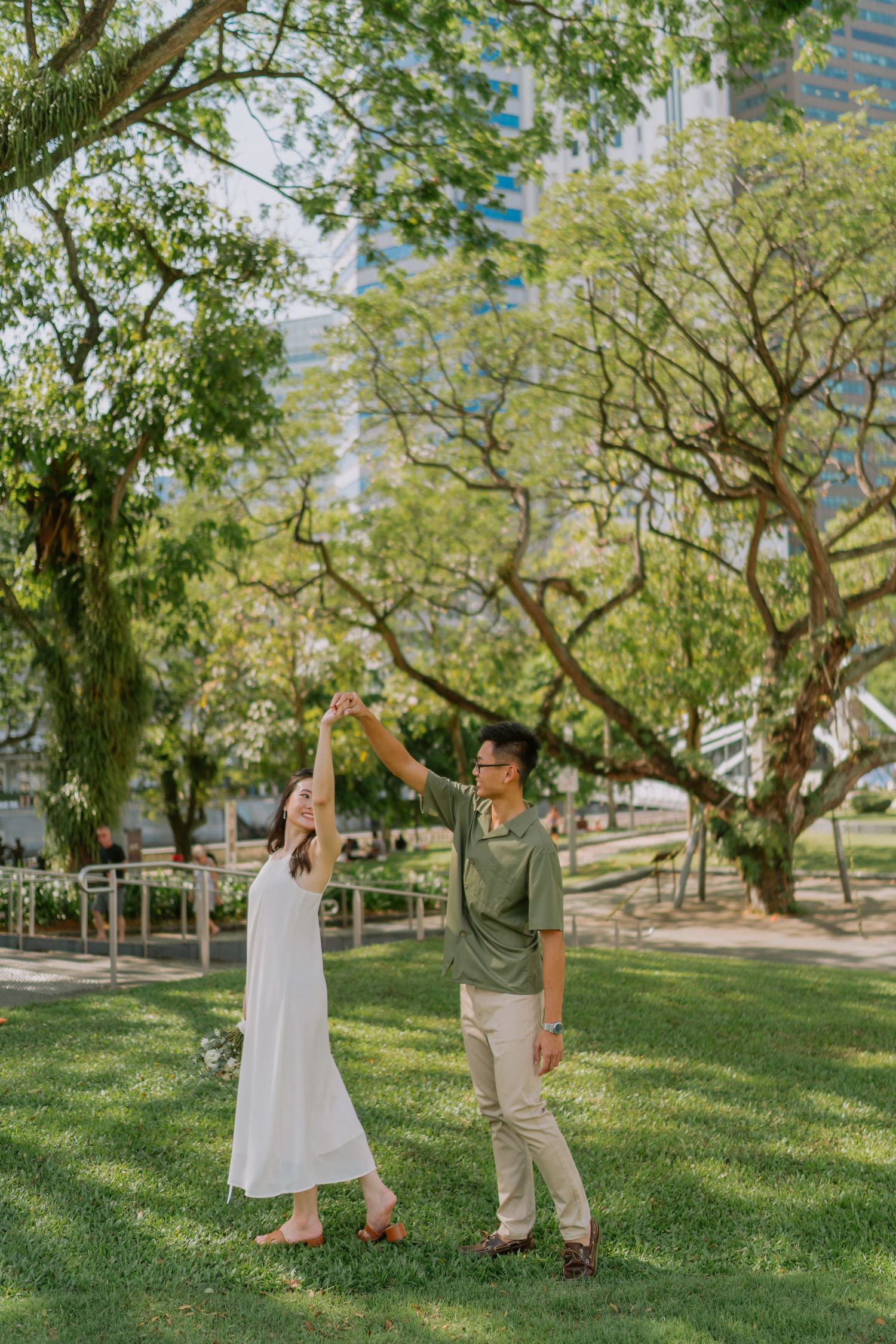 Man spinning woman in white dress under green trees in a city park.