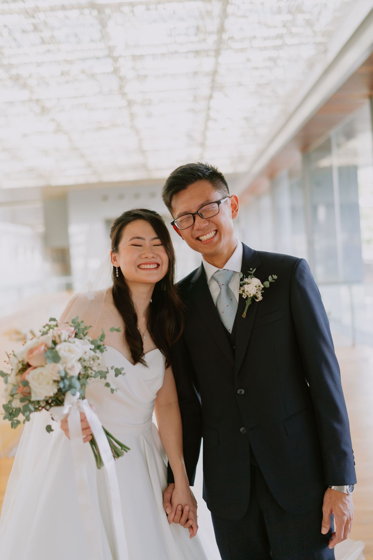 Happy Asian bride and groom laugh together holding hands inside bright modern wedding venue.