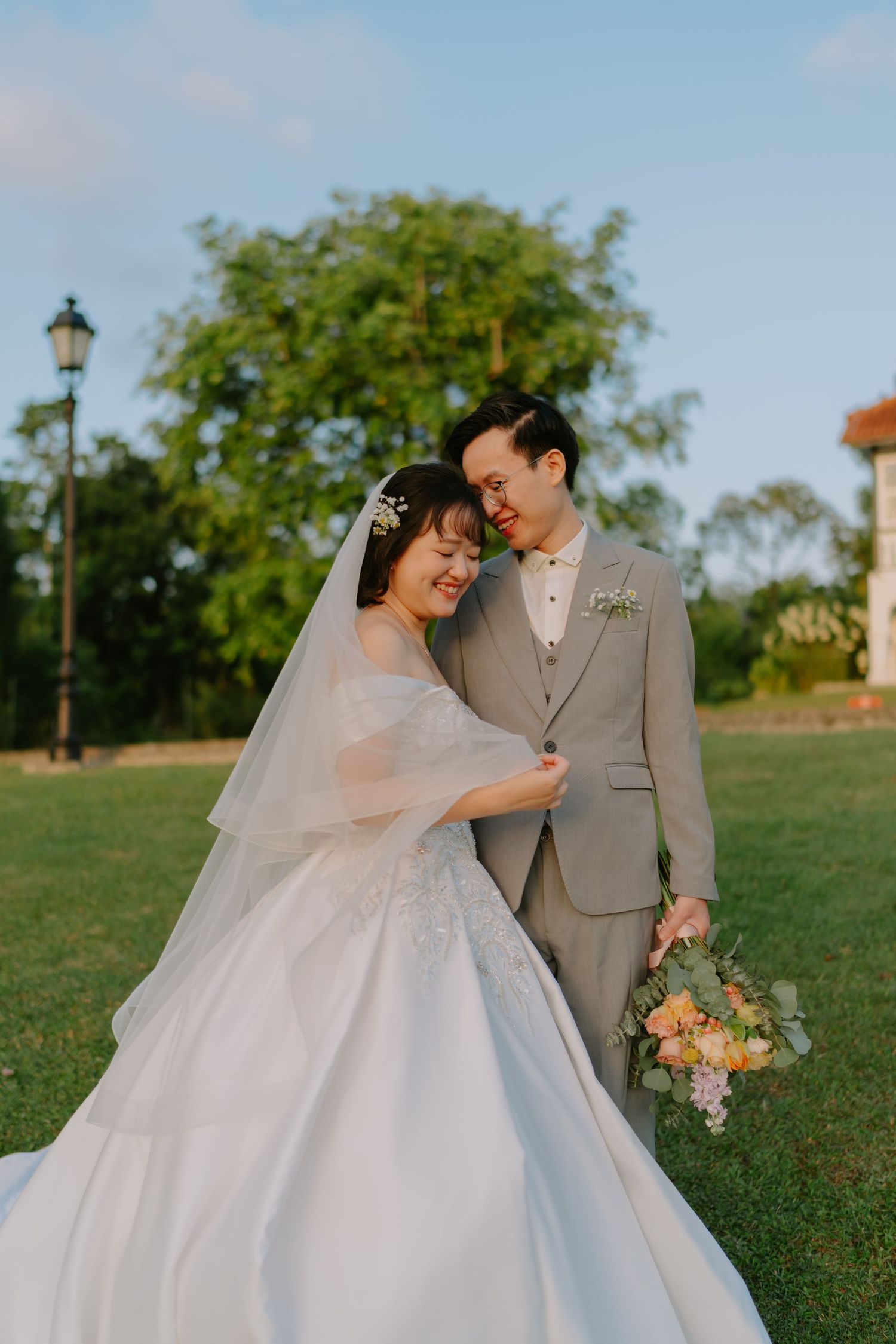 Asian couple in wedding attire sharing a tender moment outdoors, bride in white ballgown with veil.
