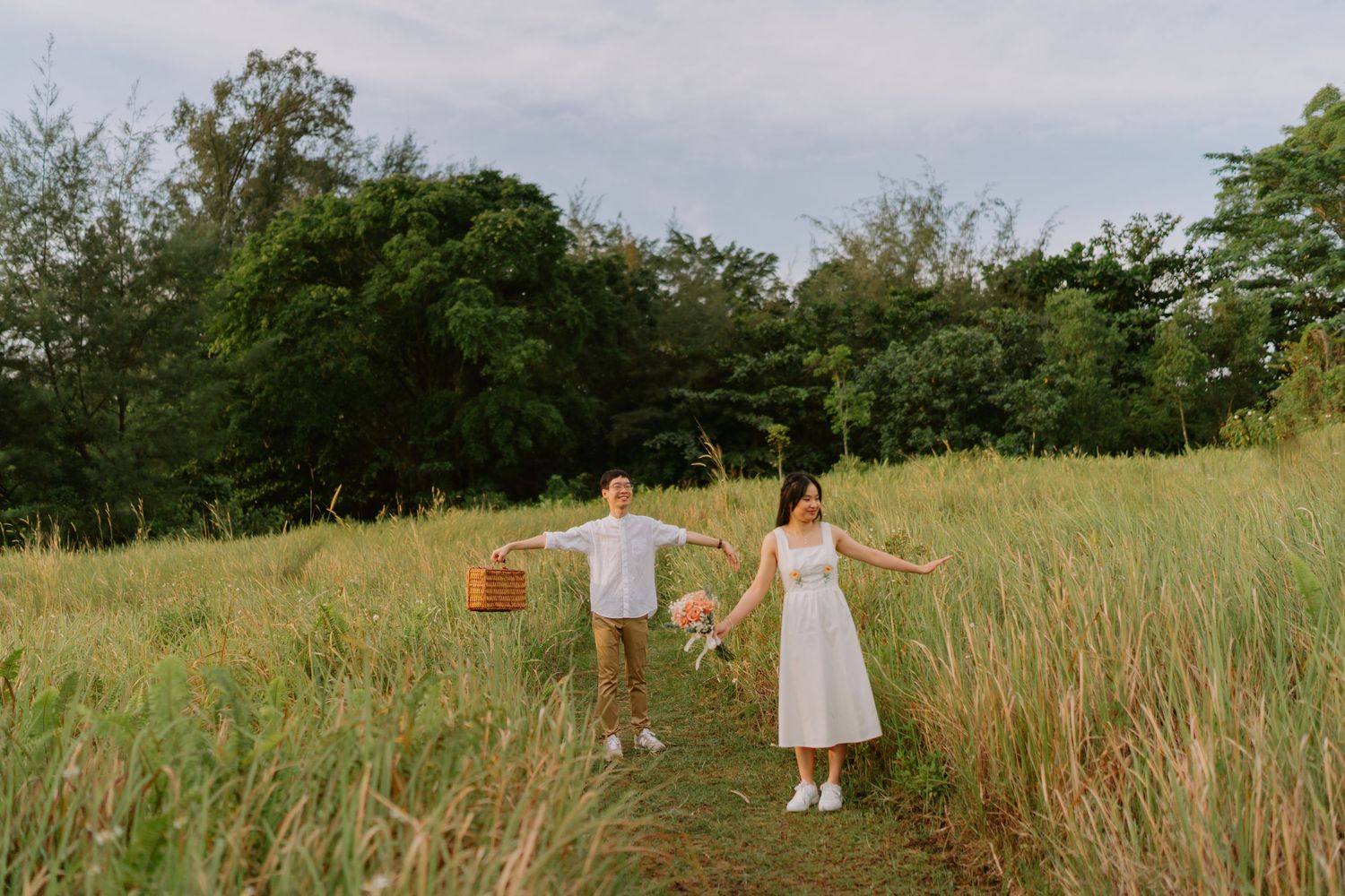 Couple playfully walking through tall grass field at dusk, arms outstretched, carrying picnic basket.