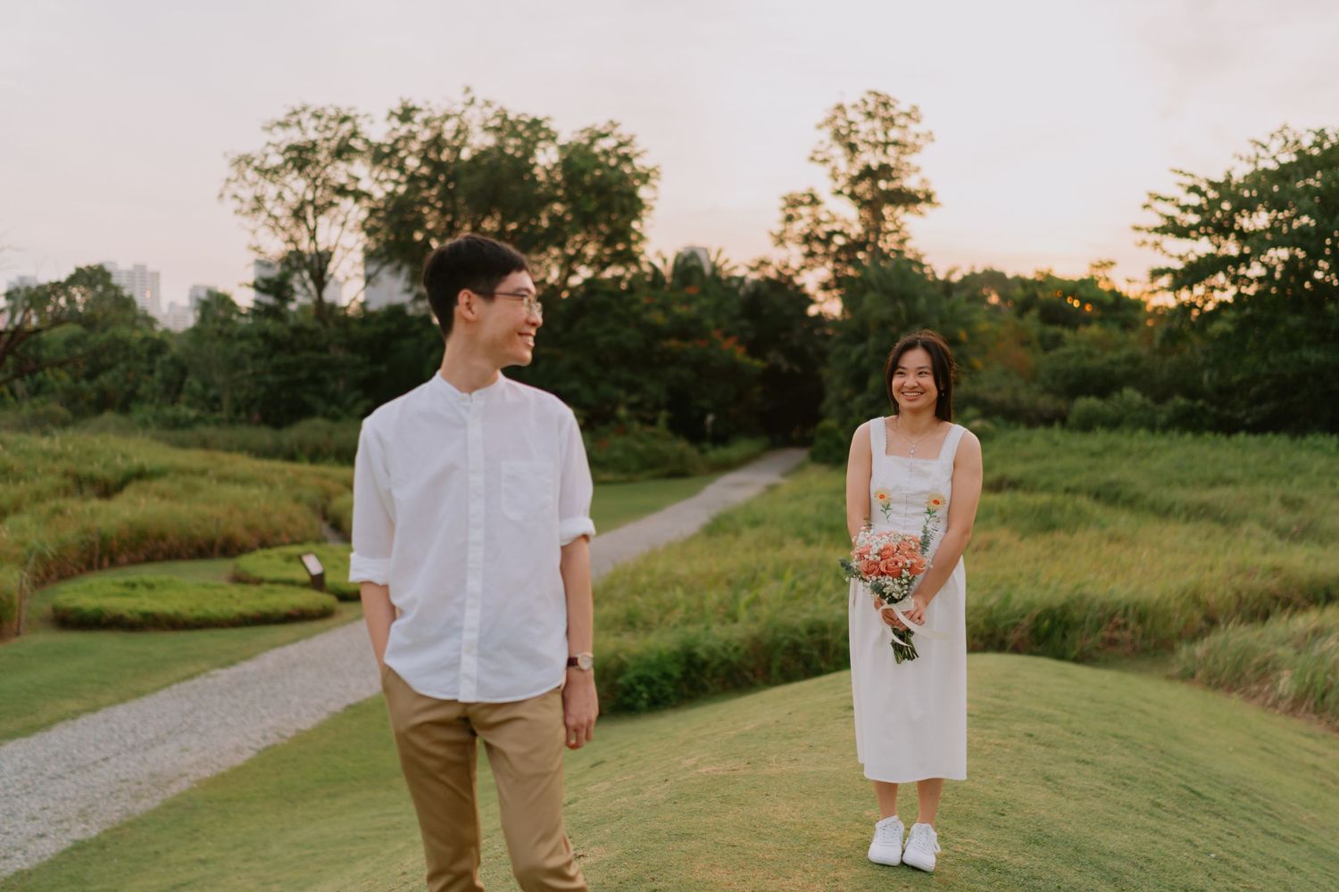 A couple stands apart on a grassy hill at sunset, the woman holding a bouquet of pink flowers.