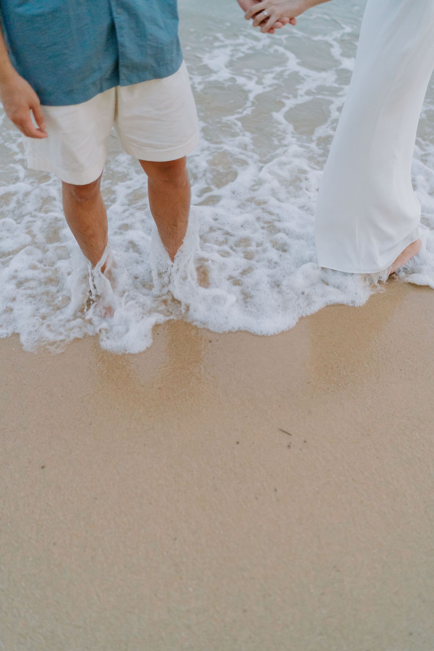 Couple standing barefoot at the shoreline, waves washing over their feet on a sandy beach.