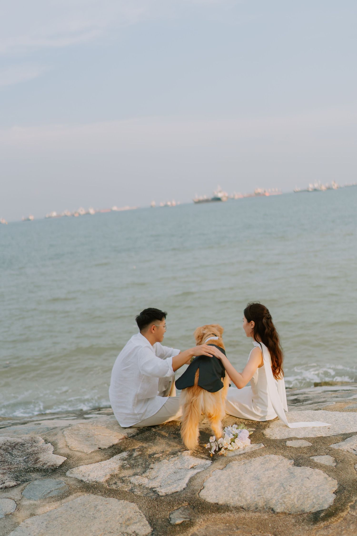Couple sitting on rocky shore with golden retriever gazing at calm ocean at dusk.
