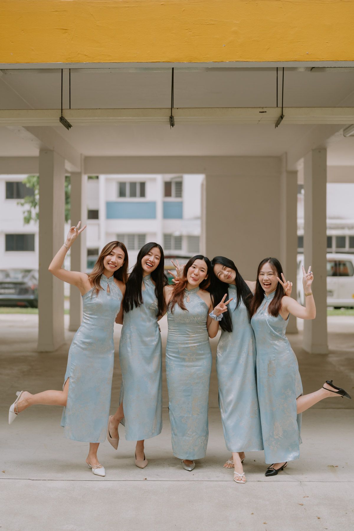 Five women in matching light blue midi dresses pose joyfully under a covered walkway, smiling and making fun gestures.
