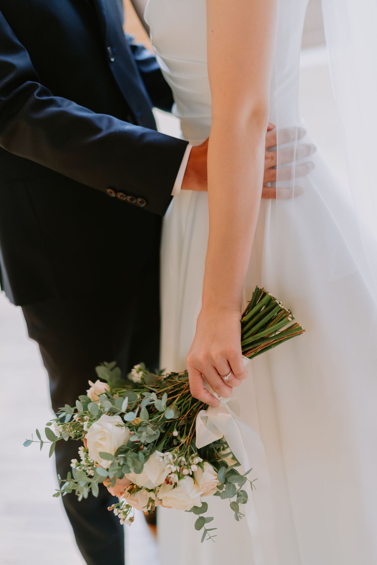 Close-up of bride holding elegant white and pink peony bouquet with eucalyptus while groom embraces her waist.
