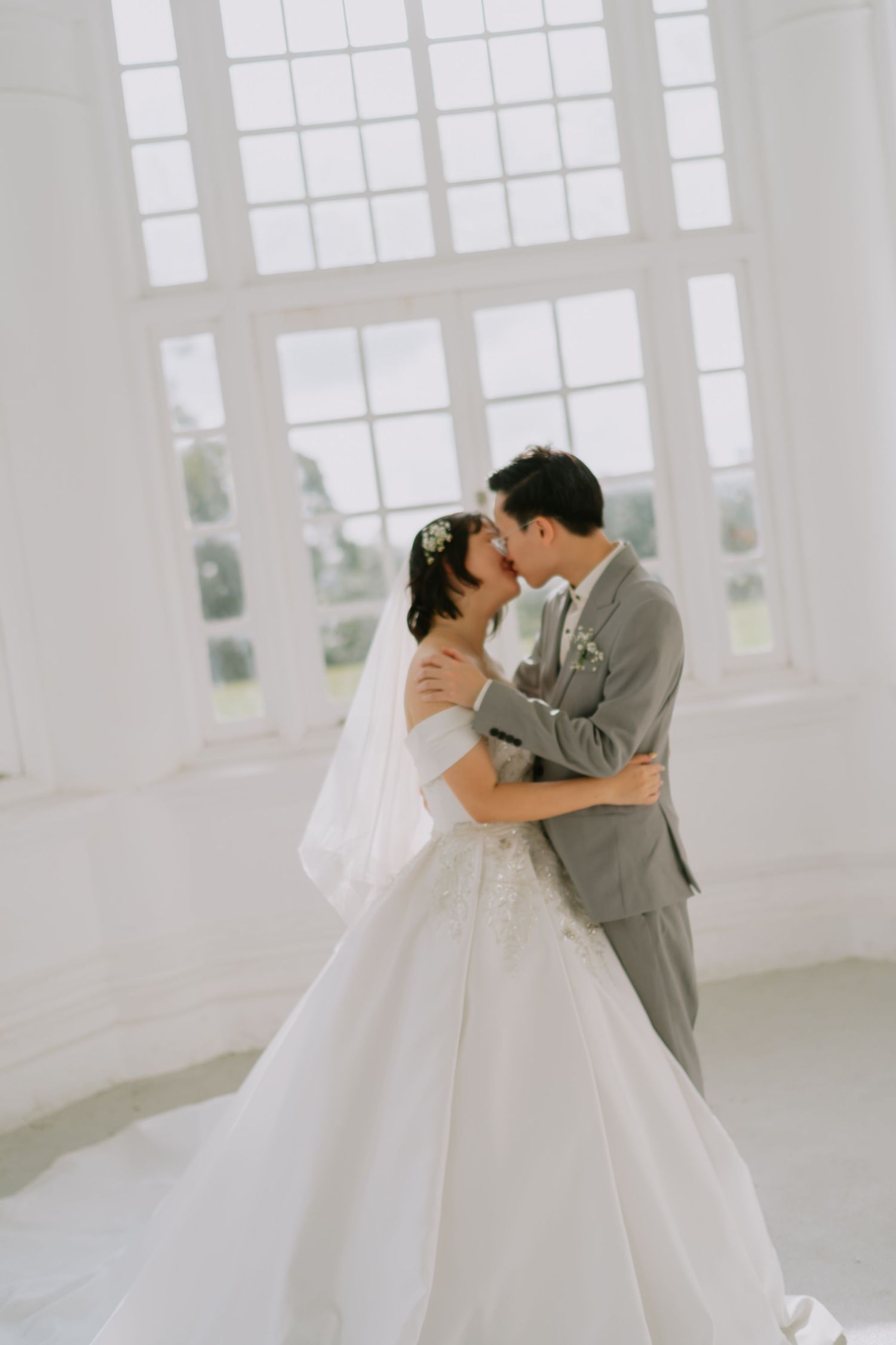 Bride and groom sharing a kiss in a bright white room with large windows on their wedding day.