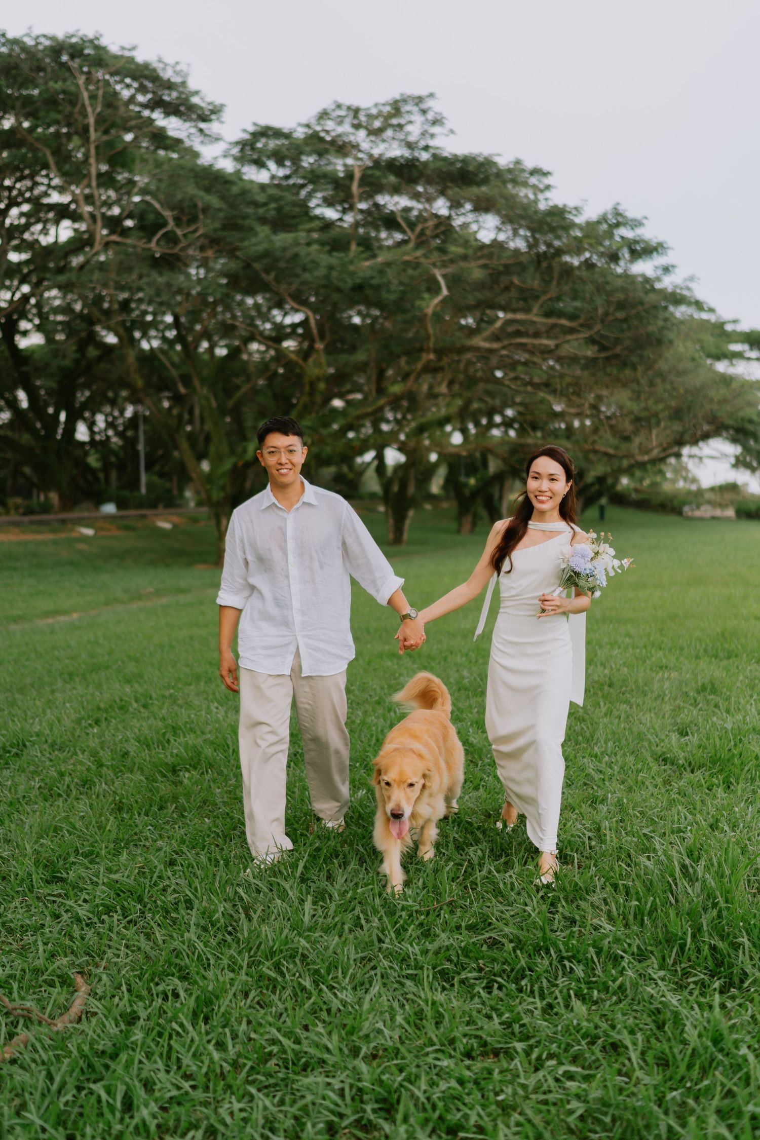 Smiling couple in white wedding outfits walk hand-in-hand with golden retriever on green grass.