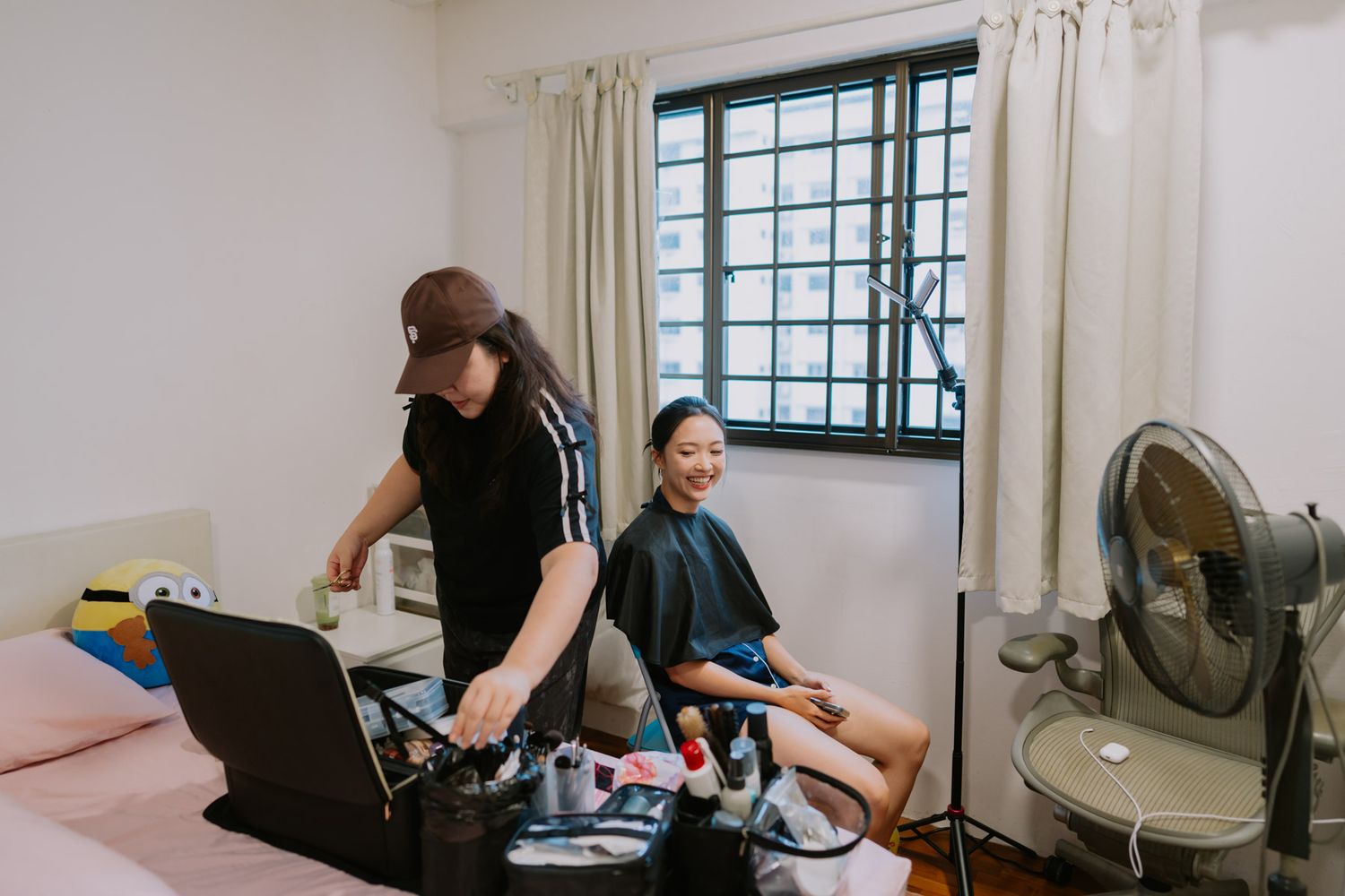 A hairstylist works on a smiling woman seated in a bedroom salon chair with makeup tools on a laptop bag.