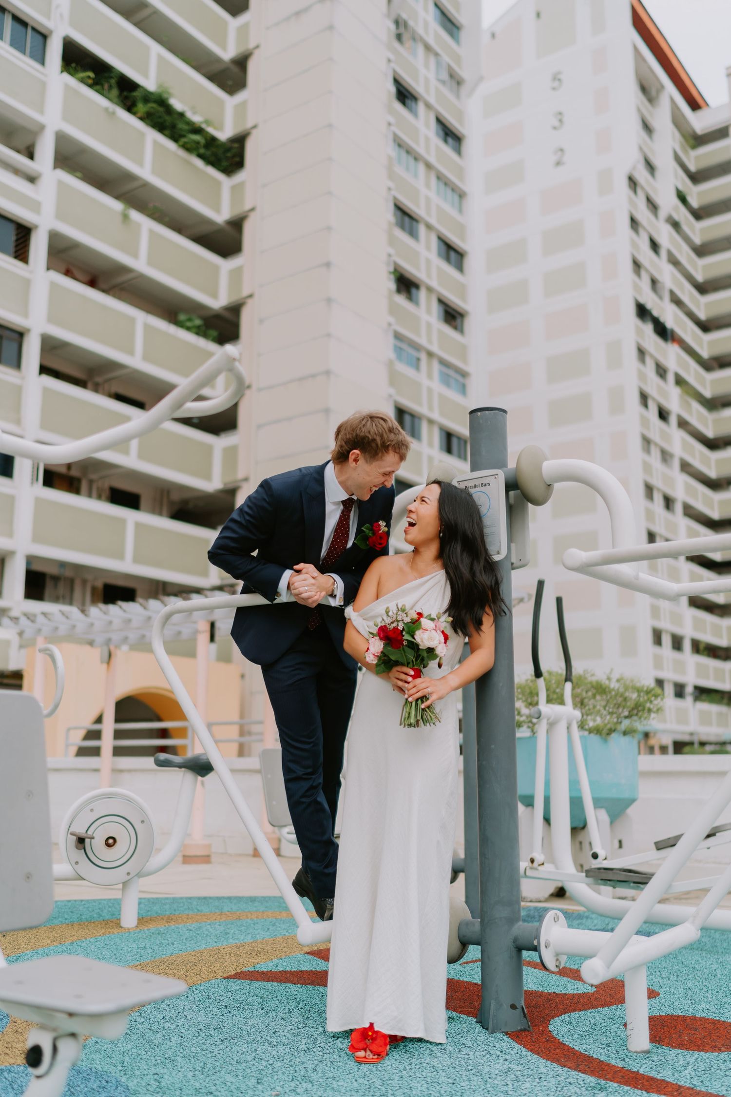Bride and groom embrace at outdoor gym equipment with high-rise apartment building backdrop.