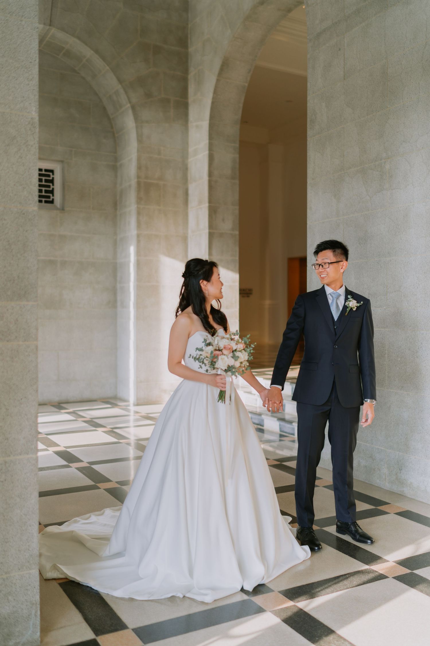 Bride in white gown and groom in navy suit hold hands on checkered marble floor in stone archway.