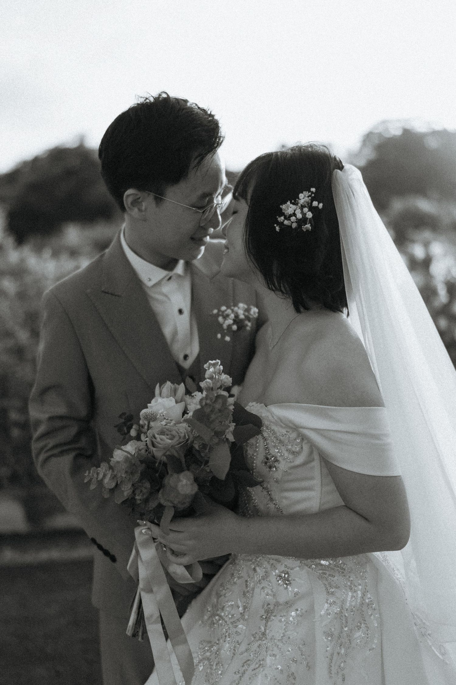 Black and white portrait of Asian newlyweds gazing at each other, bride holding floral bouquet.
