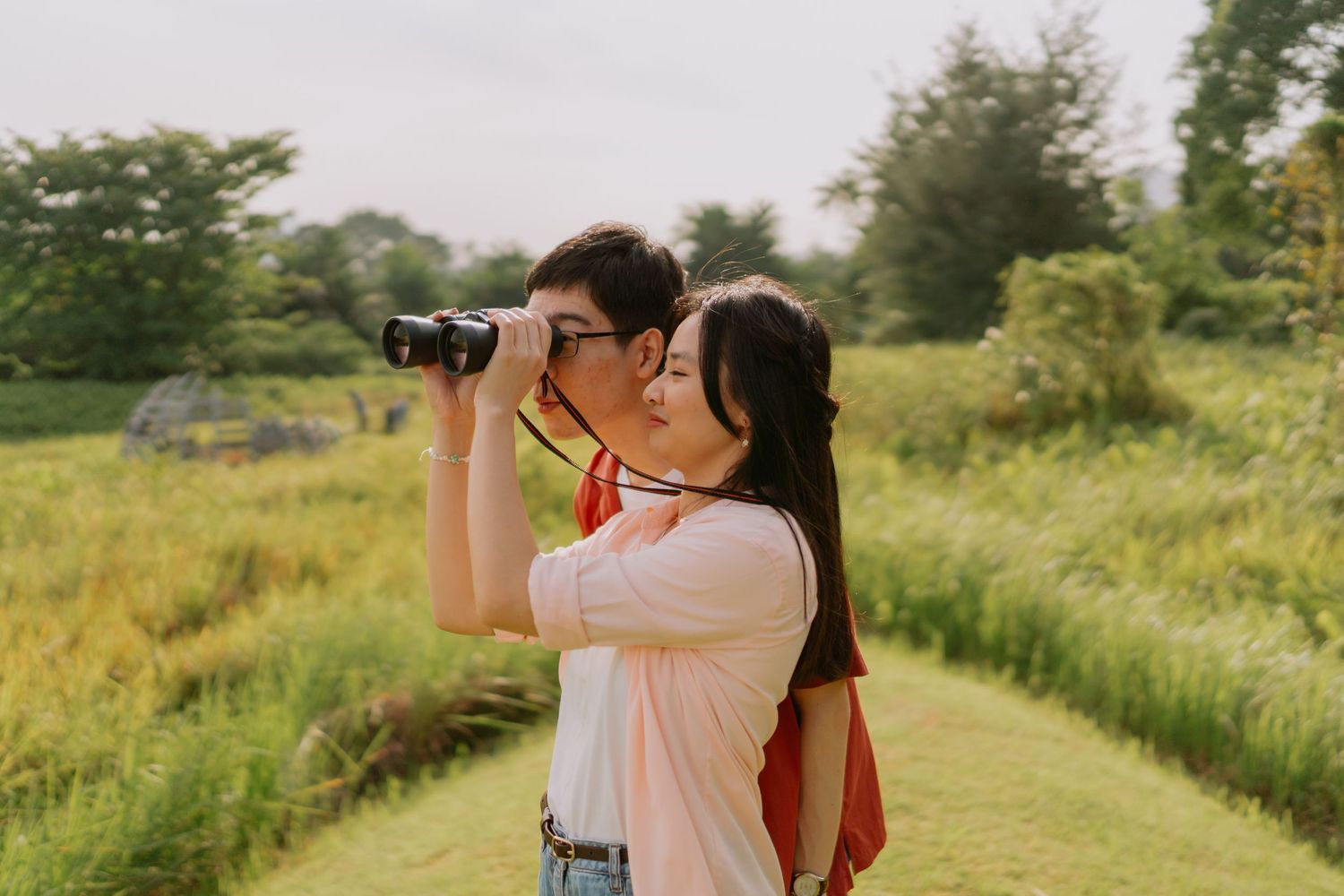 A young couple shares binoculars together while standing in a scenic green field.