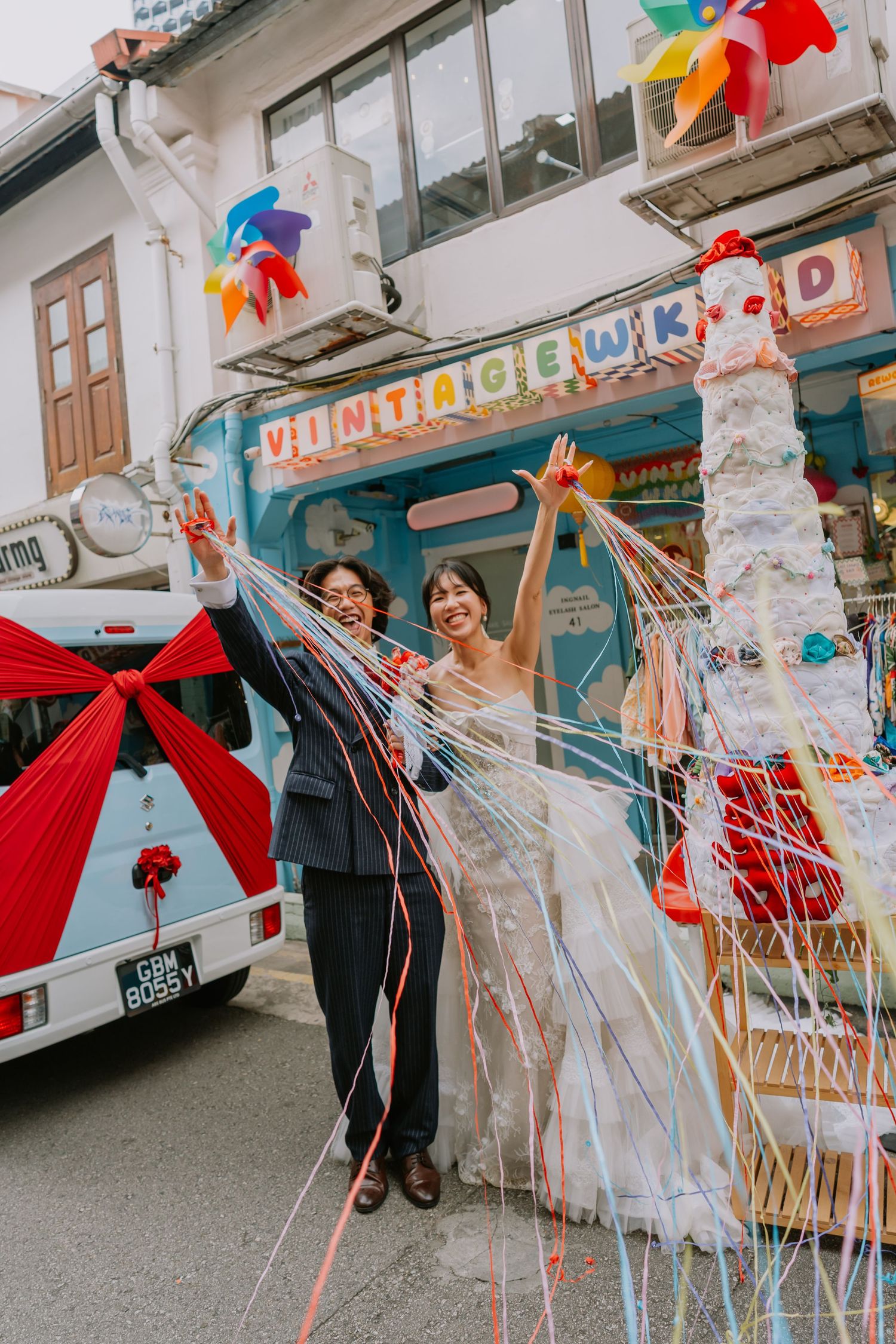 Joyful bride and groom celebrating with streamers outside vibrant Vintage Weekend shophouse.